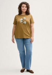 Mosterdkleurig t-shirt met een bloemendessin, gecombineerd met lichtblauwe rechttoe-rechtaan jeans en bruine platte schoenen. Zachte katoenen stof.