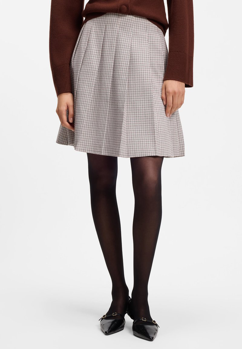 Frau trägt einen braunen Pullover, einen grauen karierten Faltenrock, schwarze Strumpfhosen und schwarze spitze Ballerinas mit goldenen Schnallen.