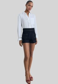 Chemise blanche en lin à manches longues avec poche et short en denim bleu taille haute avec des coutures visibles et deux poches arrière. Sandales à talons rouges.