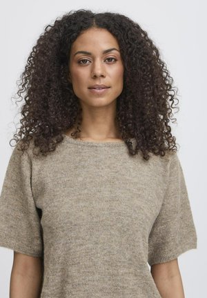 Femme aux cheveux bouclés et foncés portant un pull en tricot beige à manches courtes sur un fond clair uni.
