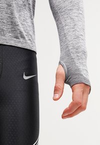 Grijs lange mouwen sporttop met duimgaten, gecombineerd met zwarte leggings met een gestructureerd patroon en het witte Nike-logo.