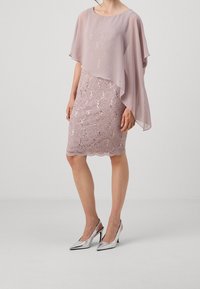 Femme portant une robe en dentelle mauve jusqu'au genou avec un superposé asymétrique transparent et des escarpins argentés à bride arrière.