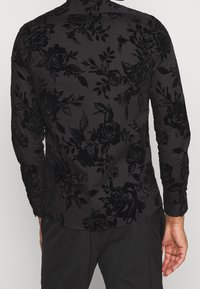 Zwarte lange mouwen shirt met een gestructureerd bloemmotief. Het materiaal lijkt zacht met een fluwelen afwerking; afgeronde zoom en knoopmanchetten.