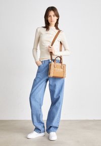 Beige lange mouwen top met geborduurd logo, gecombineerd met lichtblauwe wijde spijkerbroek en een kleine bruine handtas met bontvoering en logo. Witte sneakers.