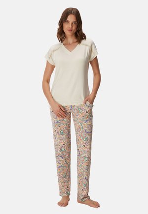 Vrouw met een beige V-hals top met korte mouwen en kleurrijke broeken met paisley-patroon, staand op blote voeten met handen in de zakken.