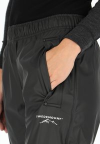 Zwarte waterdichte broek met een elastische tailleband, voorzien van een zijzak en een wit "SWEDEMOUNT" logo met berg graphics.