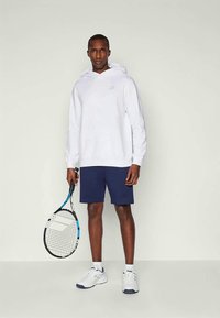 Hvit hettegenser med logo, marineblå shorts og hvite joggesko. Holder en tenniskølle med blå og svart design. Nøytral bakgrunn.