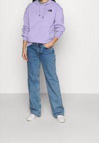 Felpa viola con tasca frontale e logo nero, abbinata a jeans a gamba larga blu e sneakers bianche. Tessuto morbido, design casual.