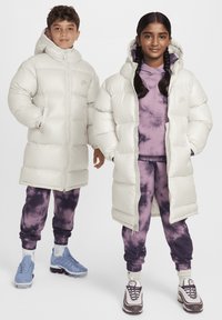 Dos niños están de pie uno al lado del otro, vistiendo ropa de abrigo. El niño lleva puesto un abrigo tipo plumas de color crema; la niña lleva un abrigo tipo plumas de color crema a juego sobre una sudadera morada y pantalones de chándal con estampados.