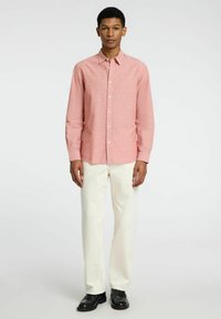 Camisa de cuadros rojos con botones y mangas largas, combinada con pantalones blancos y zapatos negros, de pie contra un fondo claro.