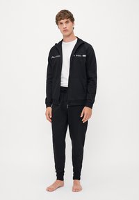 Zwarte zip-up hoodie met reflecterende accenten en logo, gecombineerd met zwarte sweatpants, beide gemaakt van zachte stof. Model staat op blote voeten.
