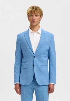 Mann mit kurzen blonden Haaren, der einen hellblauen Anzug und ein weißes Hemd trägt, steht vor einem schlichten hellen Hintergrund und schaut nach vorne.