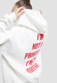 Eine Person, die die Kapuze eines weißen Sweatshirts mit rotem Text, der auf dem Rücken steht "ICH BIN KEINE PRINZESSIN, ICH BIN DIE KÖNIGIN", anpasst.