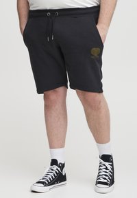 Black cotton shorts with a drawstring waist and side pockets; features a gold heart graphic on the left thigh. Paired with black high-top sneakers.
