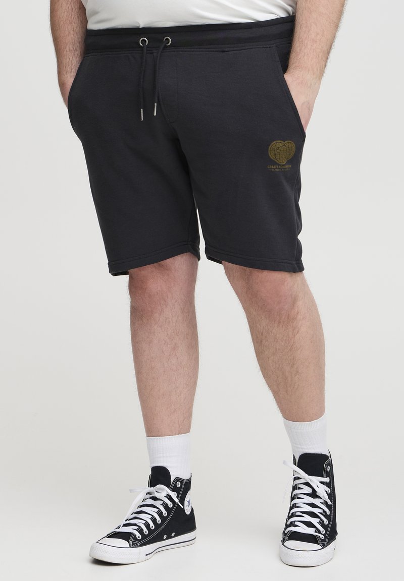 Black cotton shorts with a drawstring waist and side pockets; features a gold heart graphic on the left thigh. Paired with black high-top sneakers.