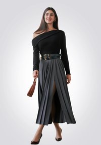 Top nero scollo off-the-shoulder abbinato a una gonna lunga grigia plissettata con uno spacco alto sulla coscia. Gli accessori includono una cintura verde e una borsa marrone.