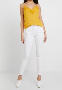 Camisole en satin jaune avec des détails en dentelle au niveau du col, associé à un jean blanc ajusté et des chaussures à talons blanches sur un fond neutre.