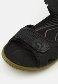 Mustad mustrik rooliga ja tekstuuriga pinnaga. Pehme jalatsipadi diskreetsete kontuuridega. Oliivroheline kummist välistald. Brändi logo rihmal.