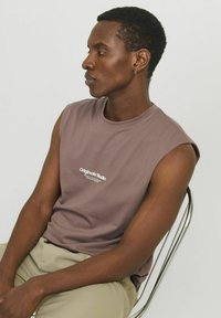 Camiseta de algodón sin mangas en color malva con un escote redondo y un logo blanco que dice "Originals Studio" impreso en el frente.