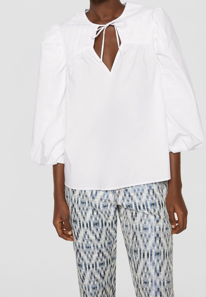 Blusa bianca con maniche a sbuffo e scollatura a chiave, abbinata a pantaloni chiari blu e bianchi con motivi geometrici.