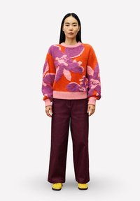 Vrouw in een feloranje en paarse gebloemde trui, met een wijde paarse broek en gele schoenen, staand tegen een effen achtergrond.