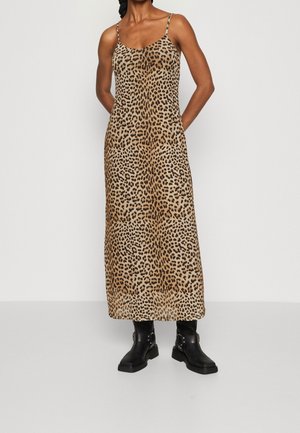 Person trägt ein langes, ärmelloses Kleid mit Leopardenmuster in Kombination mit schwarzen Stiefeletten, steht mit Händen hinter dem Rücken vor einfarbigem Hintergrund.