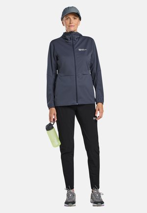 Woman in navy jacket, black pants, and cap holding a green water bottle, wearing gray hiking shoes, standing against white background.