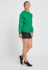 Grön hoodie med dragsko, bomullsblandat tyg, kombinerad med svarta shorts. Modellen bär svarta sneakers, neutral bakgrund.