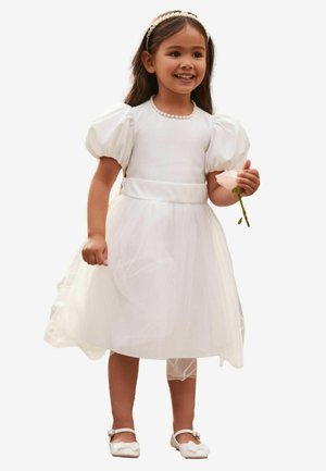 Robe en tulle blanche avec manches bouffantes, ceinture en satin et collier de perles. La fille tient une rose rose et porte des chaussures blanches avec des nœuds.