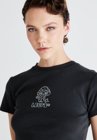Jauns cilvēks ar īsiem matiem, ģērbies melnā T-kreklā, uz kura ir zeltītas sēnes dizains un uzraksts "LEVI'S" uz gaismas fona.