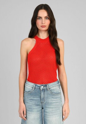 Femme aux longs cheveux foncés portant un haut rouge en tricot sans manches et un jean taille haute bleu clair, debout devant un fond gris.