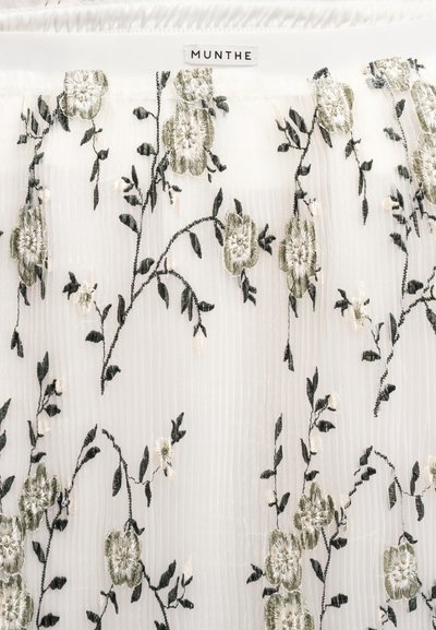 Λευκό ύφασμα με κάθετες ρίγες που διαθέτουν περίπλοκη κεντητή πράσινη και εκρού λουλουδένια διακόσμηση. Περιλαμβάνει την ετικέτα της μάρκας «MUNTHE» στο πάνω μέρος.