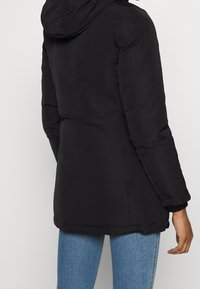 Svart puffjacka med huva, som har en mjuk yta, långa ärmar och en medellång design. Justerade muddar vid ärmarna.