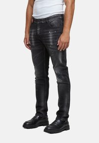 Mustat farkut slim-fit-mallilla, joissa on haalistunut design, pieniä reikiä ja valkoisia maaliroiskeita. Yhdistetty mustiin kenkiin.