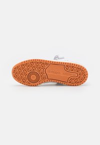 White athletic shoe sole with an orange rubber outsole featuring circular patterns and textured grip elements; brand logo visible.