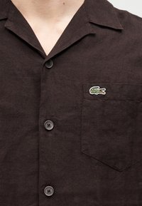 Chemise en lin marron foncé à manches courtes, dotée d'un col, d'une poche avant avec un logo crocodile brodé et de boutons sur le devant.