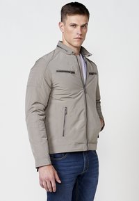Chaqueta gris de hombre con cuello alto, dos bolsillos en el pecho con cremallera, hombros texturizados y un acabado suave. Se lleva con jeans azules.