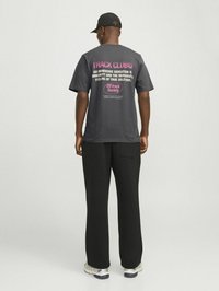T-shirt gris à manches courtes avec un texte en rose et blanc audacieux dans le dos, à coupe décontractée et en tissu doux. Porté avec un pantalon noir et des baskets.