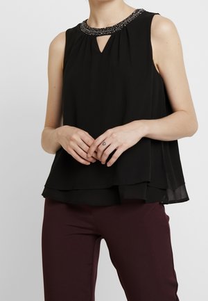 Hihaton musta pusero, jossa helmikoristeltu pääntie ja avattava yksityiskohta, yhdistetty tumman viininpunaisten housujen kanssa, mallin kädet yhteen puristettuina, sormessa sydäntatuointi.