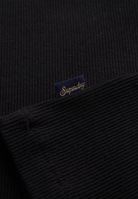 Tejido negro acanalado con una superficie texturizada. Presenta una pequeña etiqueta azul con "Superdry" en texto dorado. Vista de cerca de la costura.