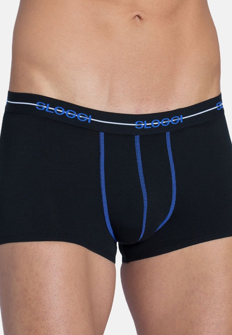 Gros plan sur le torse d'un homme portant un boxer noir avec des coutures bleues et le logo « SLOGGI » sur la ceinture, sur un fond uni.