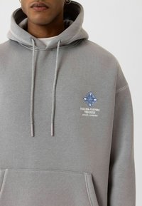Lichtgrijze hoodie met een voorzak, verstelbare koordjes, geborduurd blauw en wit ontwerp, gemaakt van zachte stof met een gladde textuur.