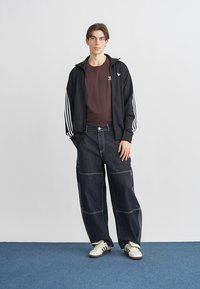 Μαύρο μπουφάν με φερμουάρ και λευκές ρίγες, καφέ μπλουζάκι και φαρδιά παντελόνια από σκουρόχρωμο τζιν. Λευκά αθλητικά παπούτσια συμπληρώνουν το ντύσιμο.