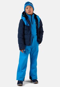 Giacca isolante blu con cappuccio sopra un top in felpa blu e pantaloni blu. Include un berretto blu con una striscia bianca. Le sneakers sono blu navy con dettagli beige.