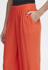 Ljusa orange vidbenade byxor med en strukturerad tyg, elastisk midja och sidofickor. Designen faller mjukt och erbjuder komfort.
