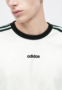 Valge Adidas t-särk musta soonikkoelusega kraega, must logoga esiküljel ja roheliste ning mustade triipudega õlgadel, tekstuurse disainiga.