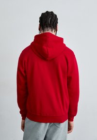 Sweat à capuche rouge avec un cordon de serrage et des poignets côtelés. Tissu doux avec une coupe ample. Porté sur un pantalon de survêtement gris, vu de dos.
