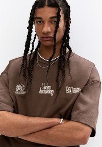 Brown cotton t-shirt featuring white text graphics: "Modern Archaeology" and logos. Short sleeves, relaxed fit, and round neckline.