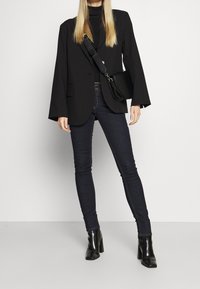 Blazer nero con maniche lunghe, abbinato a jeans scuri attillati. Stivaletti neri con tacco a blocco e una piccola borsa a tracolla nera.