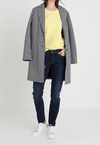 Schwarz-weißer karierten Mantel über einem gelben Hemd, kombiniert mit dunkelblauen Jeans und weißen Sneakers, präsentiert einen lässigen Lagenlook.